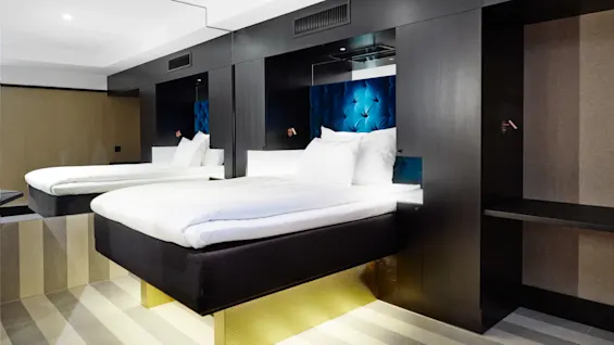 Et moderne hotelværelse har to hævede senge med hvidt sengetøj og blå tuftede hovedgærder. Sorte rammer omgiver sengene, og gulvet har stribet gulvtæppe.
