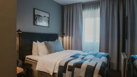 Ett inbjudande hotellrum med en säng, mönstrad filt och mjuka kuddar. Ljus strömmar in genom fönstret och skapar en lugn atmosfär.