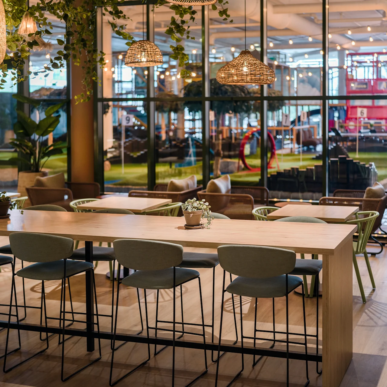 A long wooden table with chairs sits in a cozy, modern café adorned with hanging lights and plants, overlooking a bright room with grass patches and furniture through glass walls.