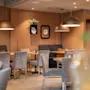Dining tables with gray chairs are arranged in a cozy, warmly-lit restaurant. Pendant lights hang above, and decorative plates adorn the brown walls, creating an inviting atmosphere.