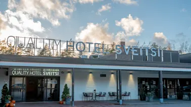 Kutsuva näkymä Quality Hotel Sveitsin ulkoasusta, jossa on ulkoistuimia ja kausikoristeita pilvisen taivaan alla.