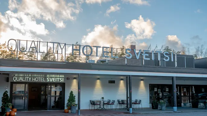 Kutsuva näkymä Quality Hotel Sveitsin ulkoasusta, jossa on ulkoistuimia ja kausikoristeita pilvisen taivaan alla.
