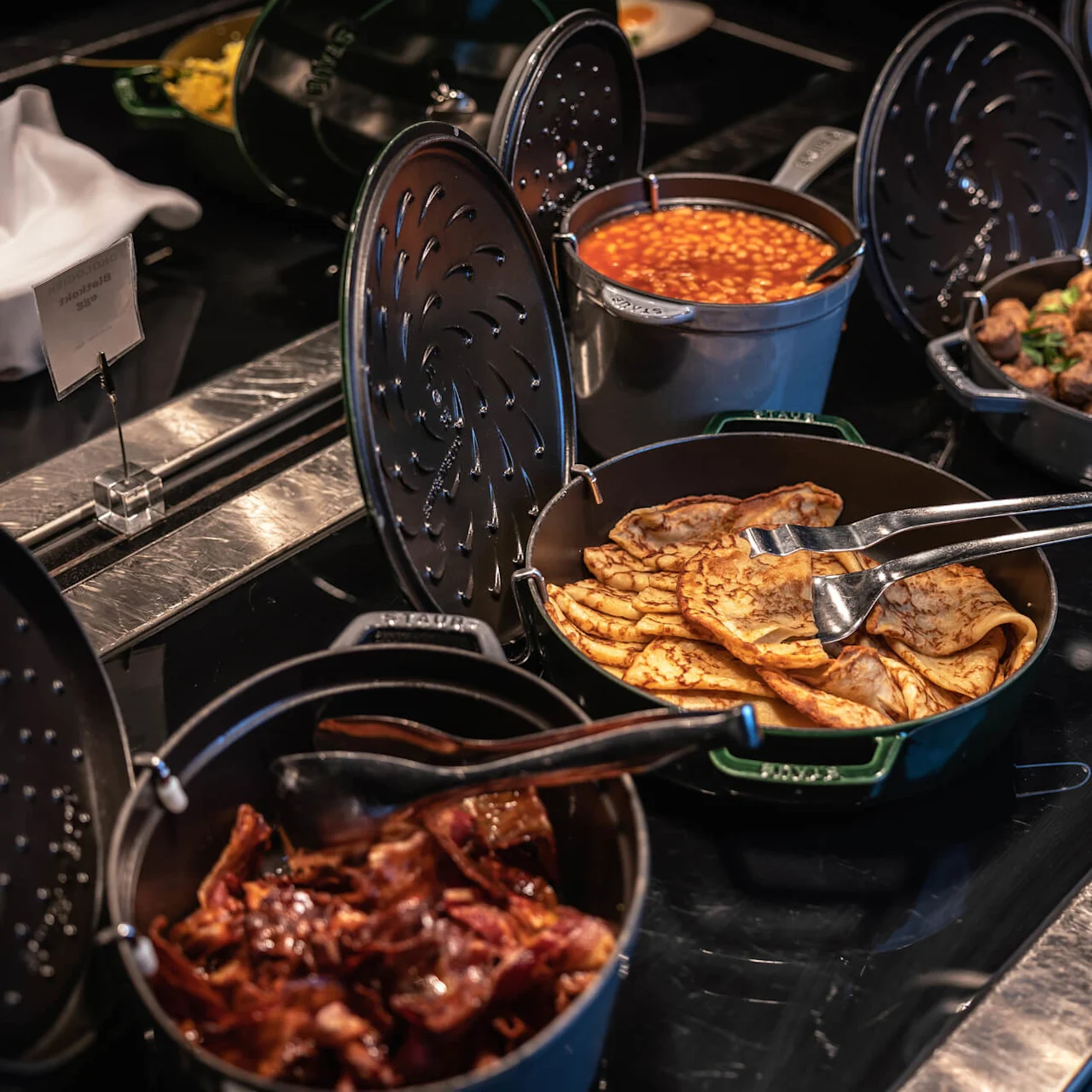 Pandekager ligger i en pande med tang på en buffet. I nærheden er bagte bønner, pølser og bacon. En dækket skål har et lille skilt med teksten "Lepeske Blinis/Crepes."