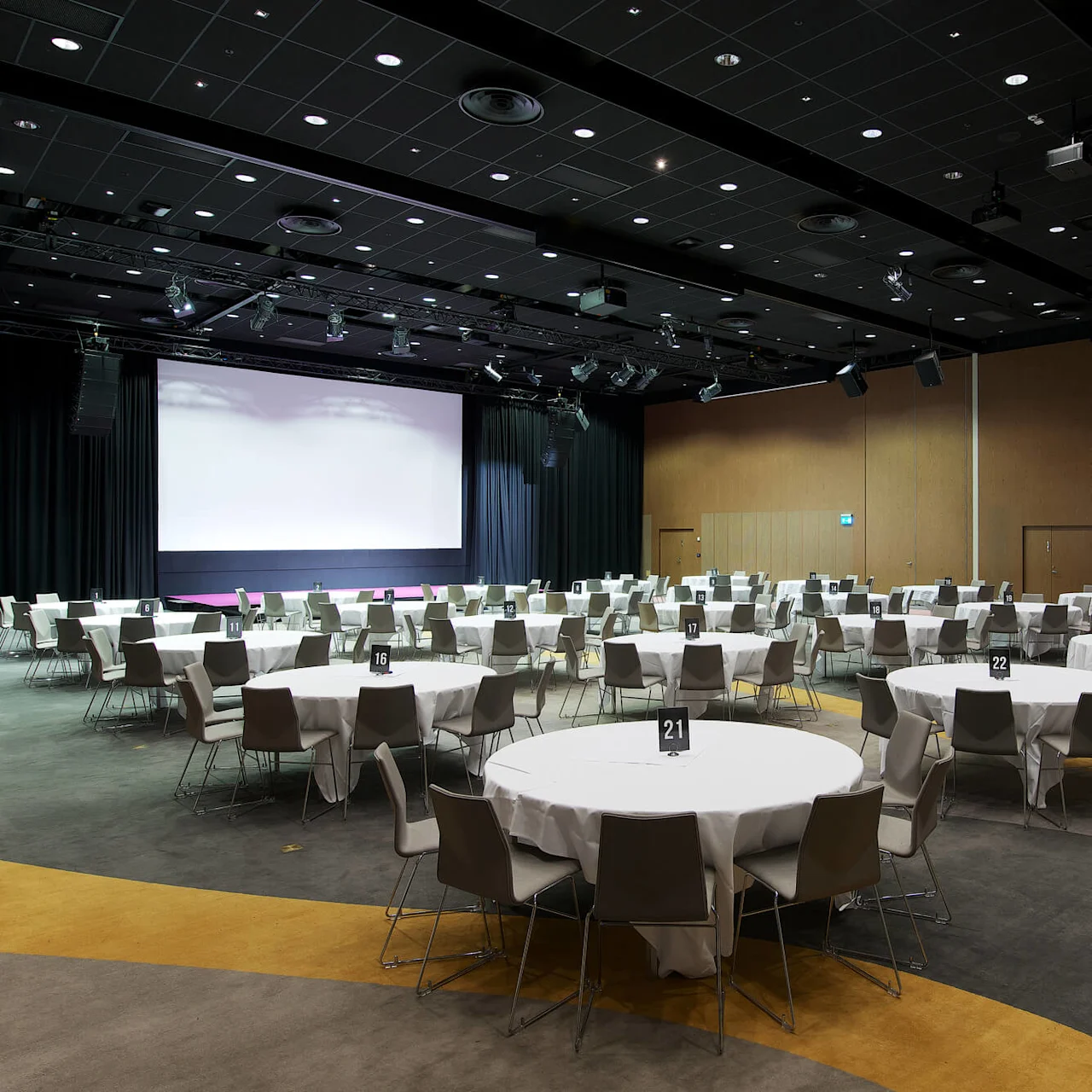 Round tables covered with white cloths, surrounded by chairs, stand in a large, carpeted conference room with a stage and a blank projector screen. Tables numbered 16, 17, 21, 22, 23.