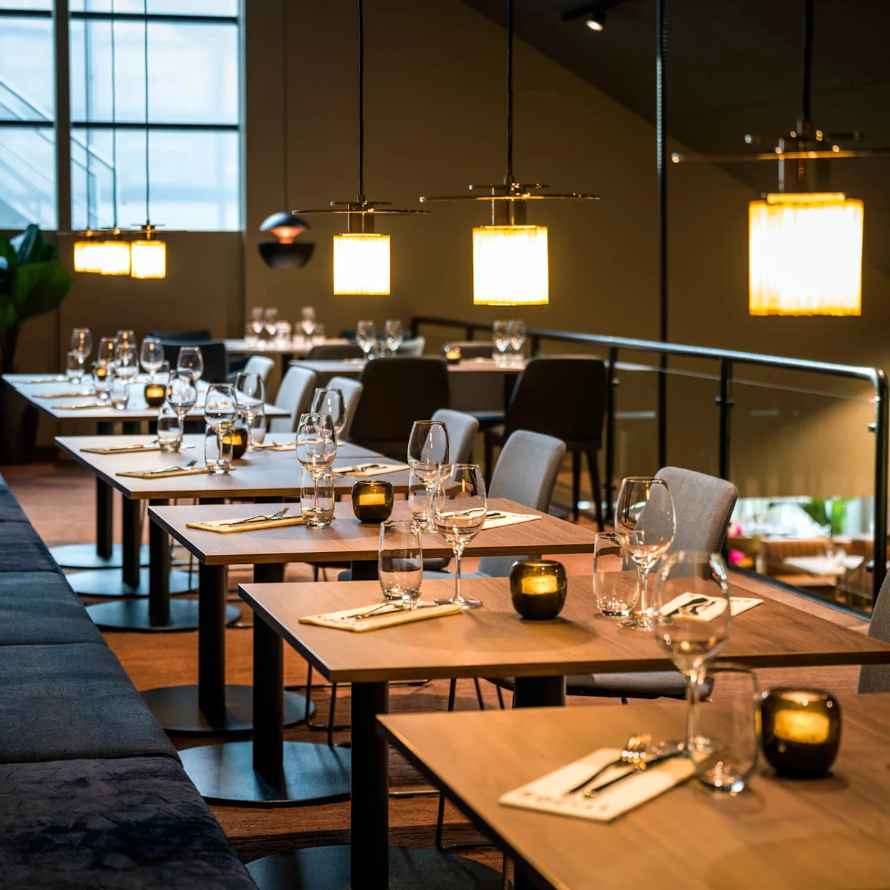 Elegante trebord med glass og brettede servietter står på rad i en koselig restaurant, mykt opplyst av hengende taklamper, med en plysjbenk langs den ene siden.
