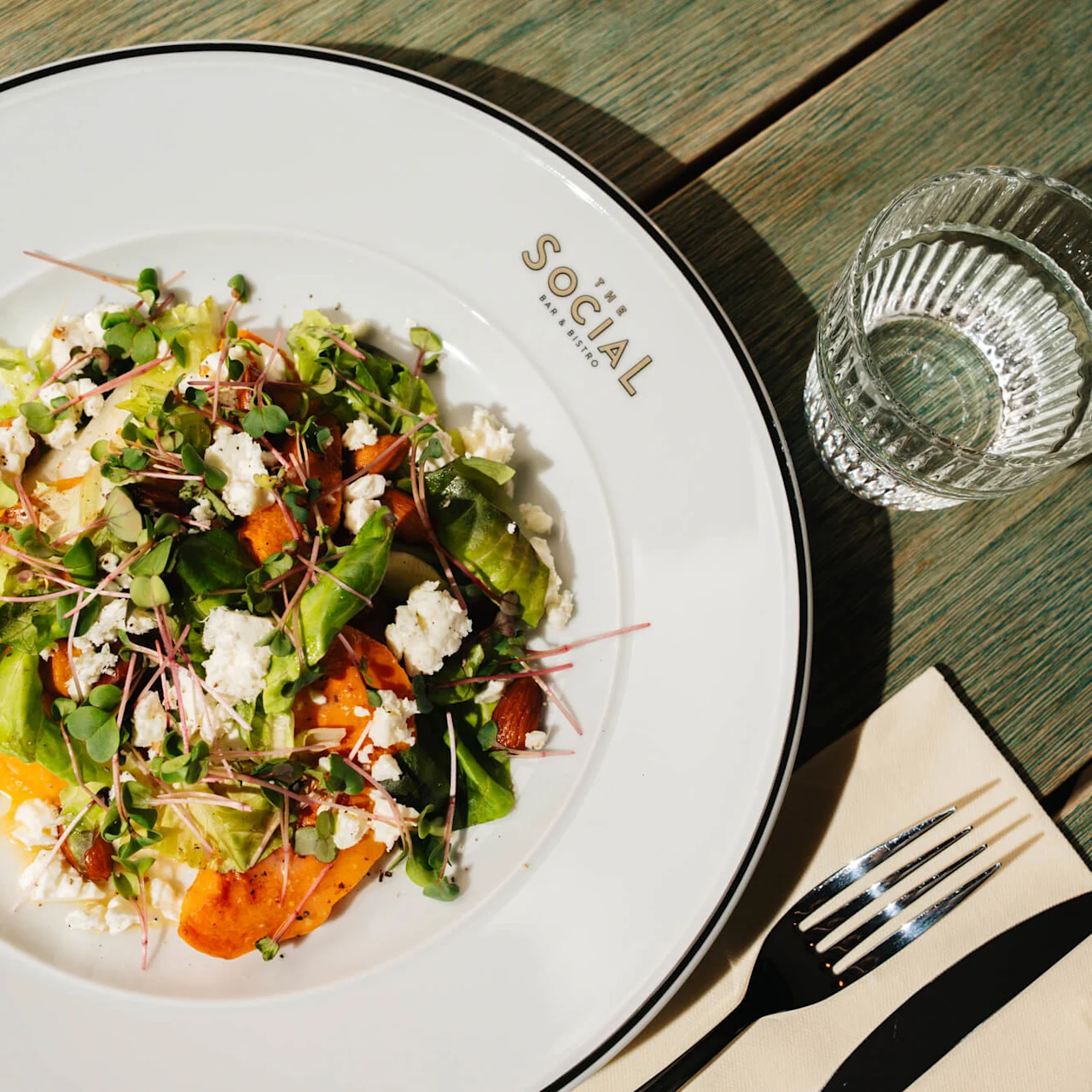 En frisk salat med grøntsaker, ost og mandler på en hvit tallerken merket "SOCIAL." På et trebord ved siden av et glass vann, gaffel, kniv og serviett.