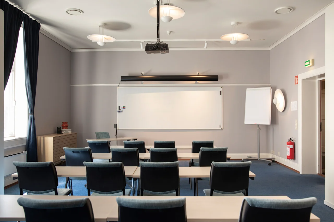 Et moderne konferencelokale med rækker af borde og stole vendt mod en whiteboardtavle og projektor, klar til en præsentation eller et møde.