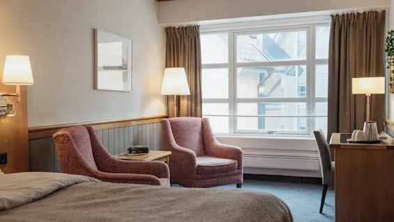 A comfortable hotel room featuring a bed, two cozy armchairs, a desk, and a large window offering natural light.