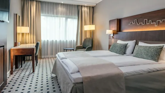 A neatly made double bed with green pillows sits in a modern hotel room. Nearby, a desk under a window with closed curtains. Warm lighting provides a cozy atmosphere.