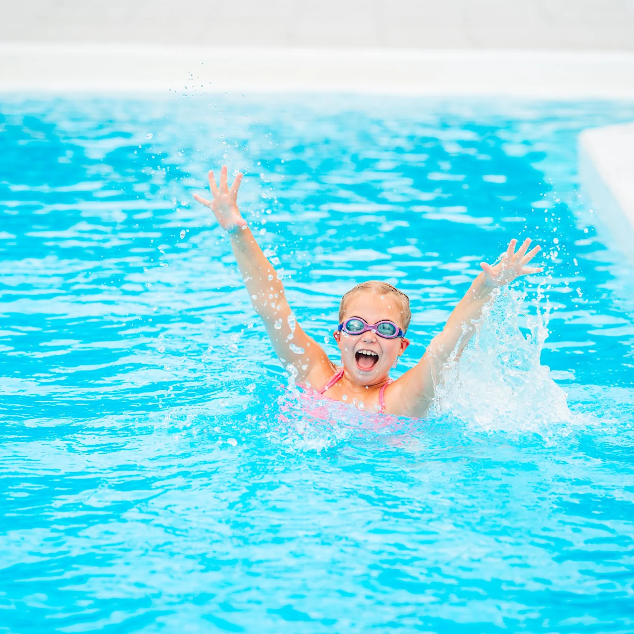 Child wearing goggles, splashing in a swimming pool with arms raised, creating water droplets around. Surrounding context includes blue water and white pool edges.