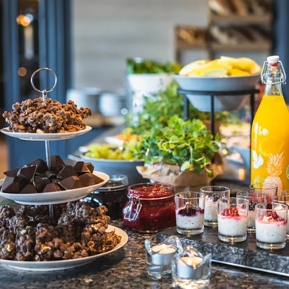 Et utvalg av desserter og frukt vises på en buffébord, med stablede tallerkener av sjokolade og bakverk, en flaske appelsinjuice og små glass med parfait.