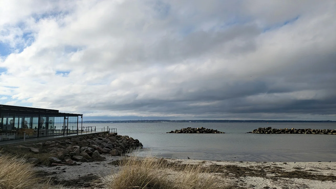 En skyet dag på stranden i Helsingborg, med en moderne bygning ved vannet og steinete moloer i sjøen.
