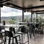 En moderne takterrasse-restaurant med høye bord og barkrakker, dekket for bespisning, med naturskjønn utsikt over grønne åser.