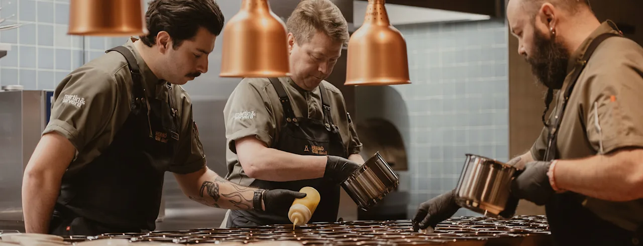 Kolme kokkia valmistaa annoksia ravintolakeittiössä, yllään yhtenäiset univormut ja hanskat, kulhoja tiskillä lämpimien riippuvalaisimien alla. Teksti univormuissa: "Marto & Girot, Xenius."