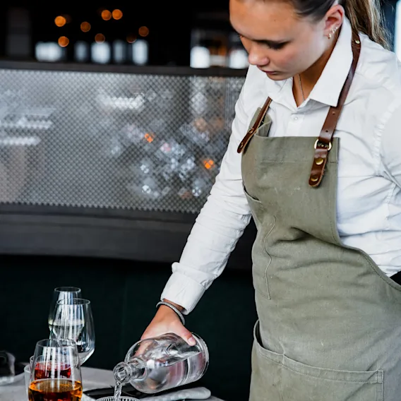 Servitrisen häller vatten från en glasflaska i ett glas vid ett bord med tallrikar, vinglas och bestick, i en svagt upplyst restaurang med hängande lampor.