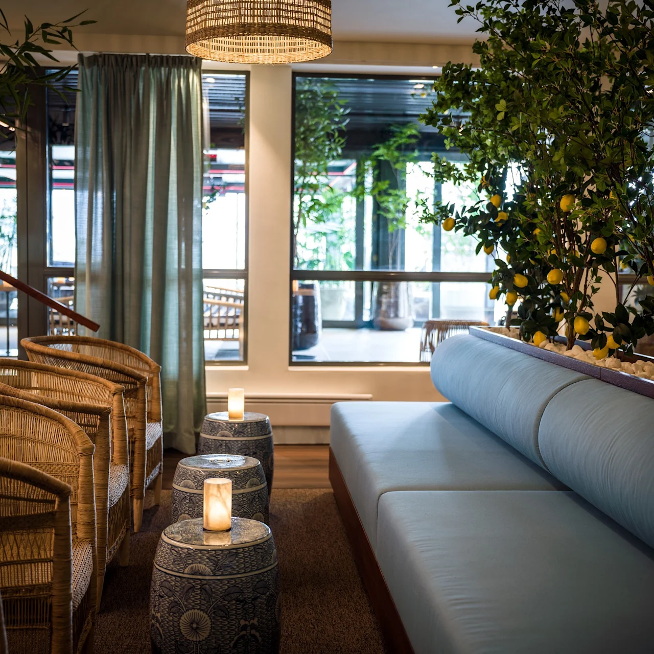 Flätade stolar och blå vadderade sittplatser är arrangerade i ett mysigt inomhusområde med krukodlade citronträd. Små bord håller tända ljus, vilket skapar en rofylld, dämpad atmosfär.