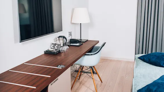 Et vægmonteret tv ved siden af et træbord med lampe, telefon, kedel, kopper og stol i et moderne rum. Tekst: “THIS IS YOUR MINIBAR”.