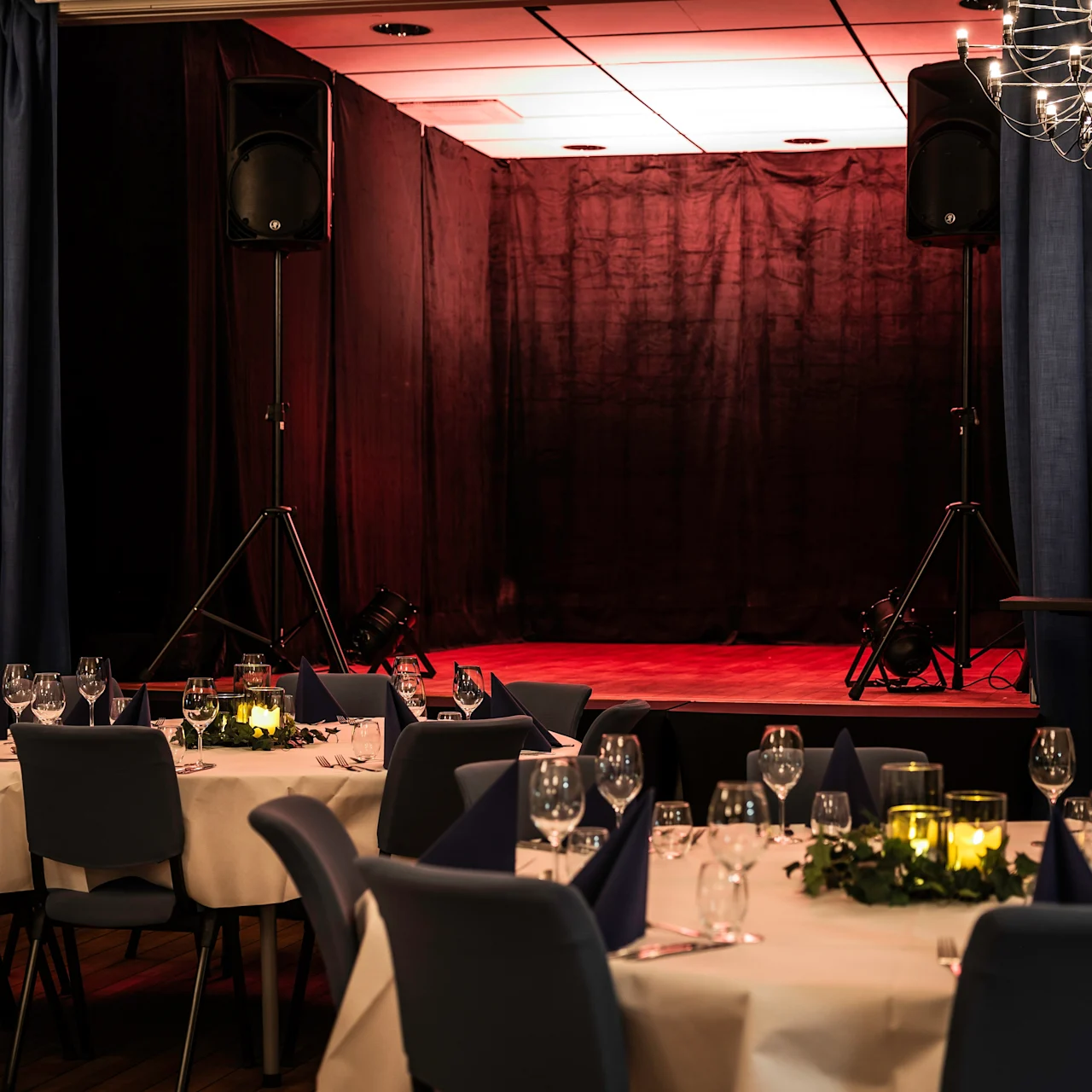 A stage with speakers is set up for a performance, surrounded by elegantly set dining tables with candles and glasses, in a dimly lit banquet hall adorned with blue curtains and chandeliers.