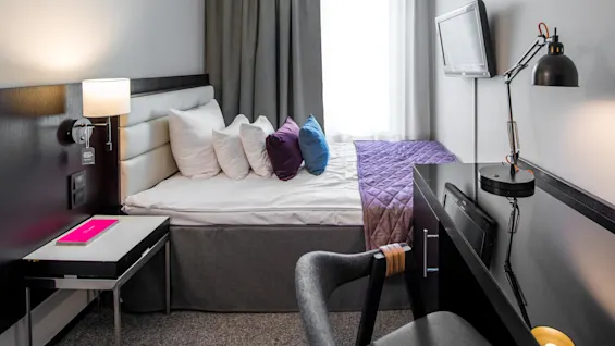 Seng med flere puter står mot en vegg i et moderne rom med skrivebord, stol og veggmontert TV.