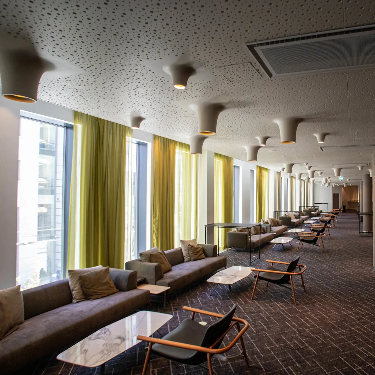 A long, modern lounge with cushioned sofas, tables, and chairs. Large windows with green curtains allow natural light. Unique ceiling lights and a carpeted floor complete the setting.