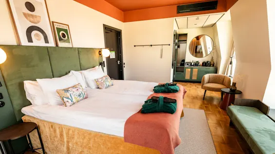 A neatly made bed with green pillows and blankets lies under framed artwork. Two green robes rest on an orange throw. A round mirror and chair are in the modern hotel room. Text: “Espresso.”
