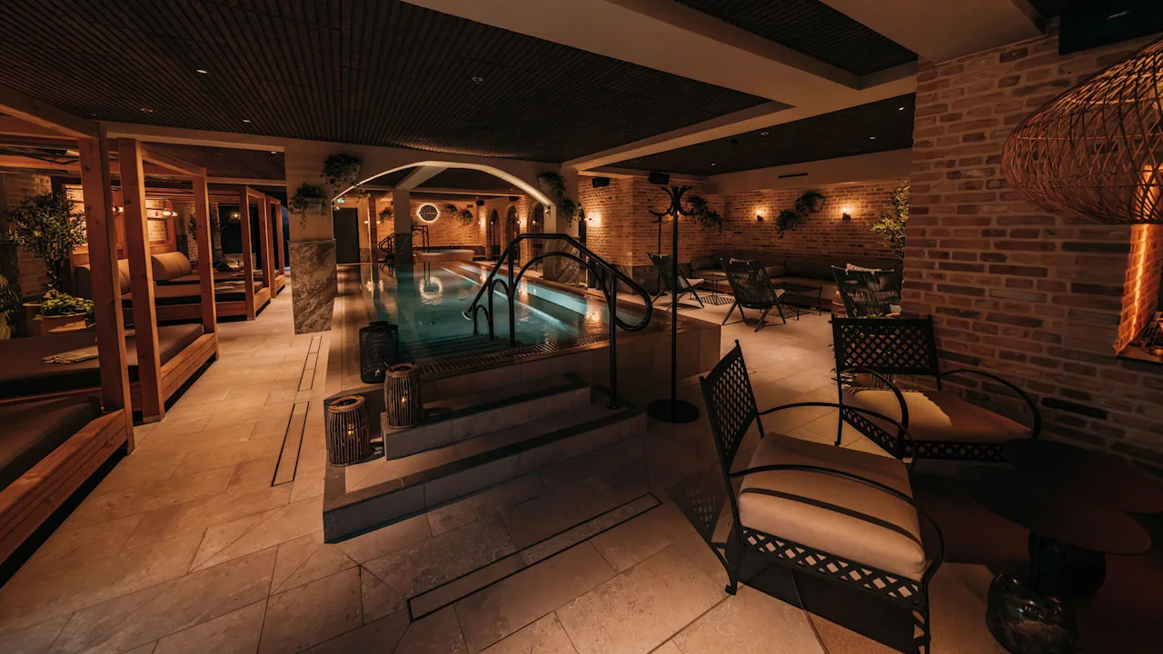 A swimming pool surrounded by lounge chairs and dim lighting in a cozy, indoor spa setting with brick walls and plants.