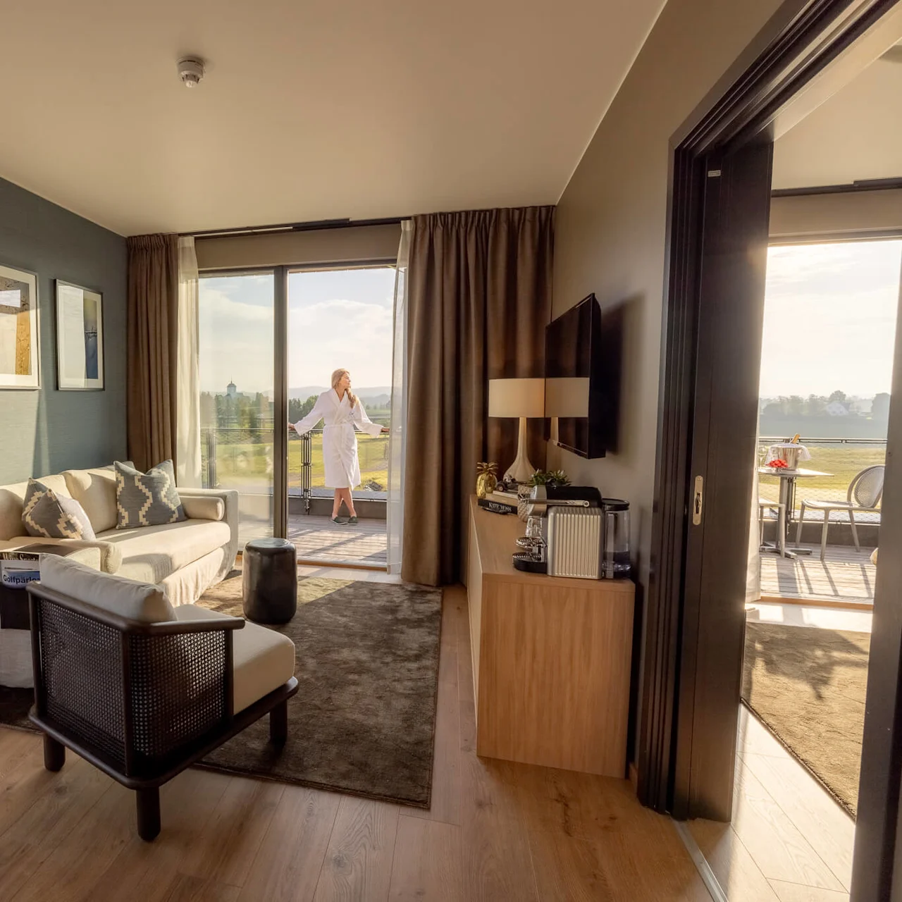En kvinna i badrock njuter av utsikten från en balkong i ett välutrustat hotellrum med modern inredning och möbler.