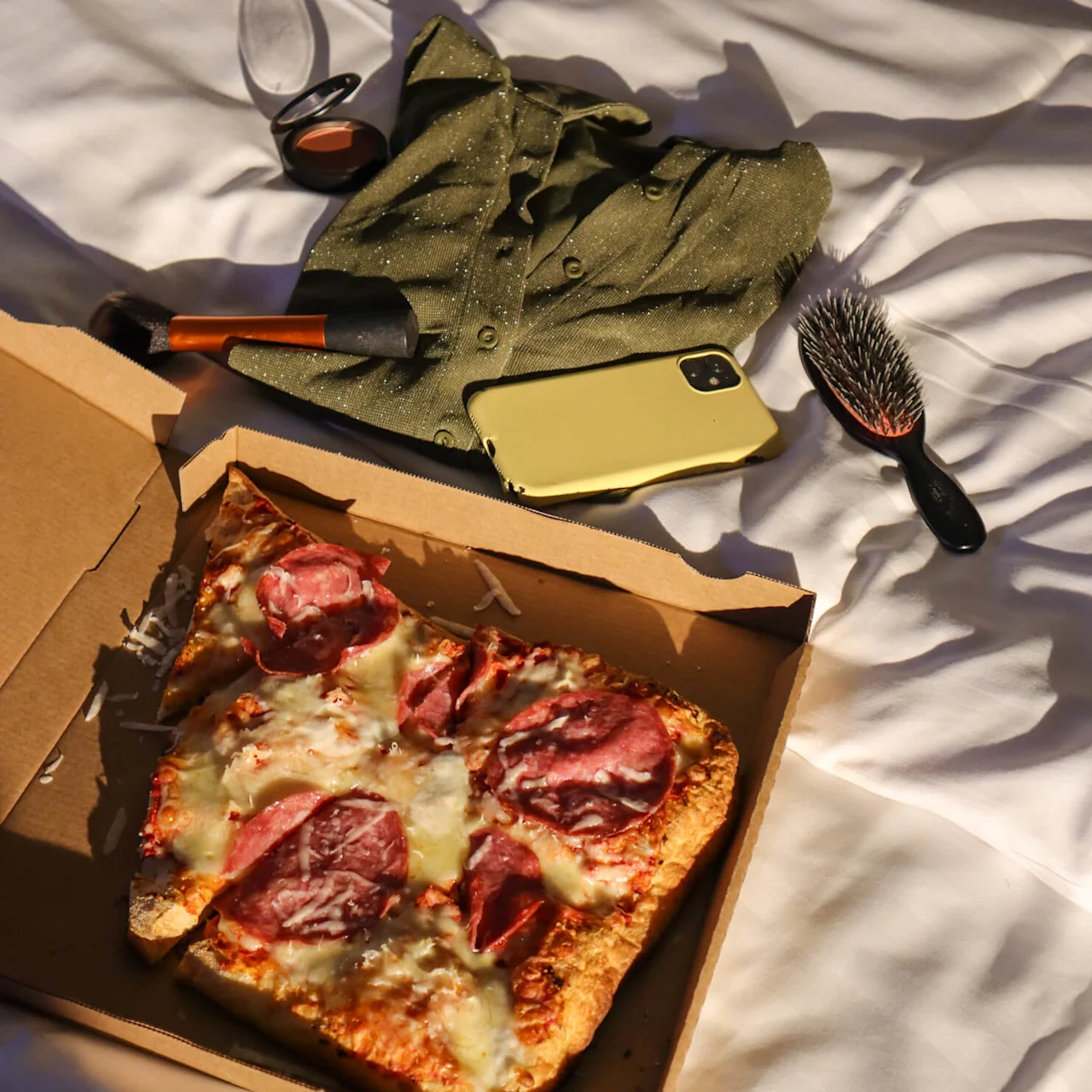 A half-eaten pizza in a box lies on a white, rumpled bedsheet. Personal items, including a green shirt, a phone, and makeup, are scattered around.