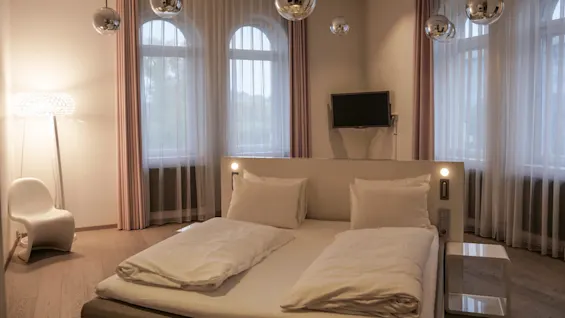 Bed with white linens sits in a modern bedroom, surrounded by floor lamps, a white chair, and hanging silver lights. Three large windows with sheer curtains provide natural light.