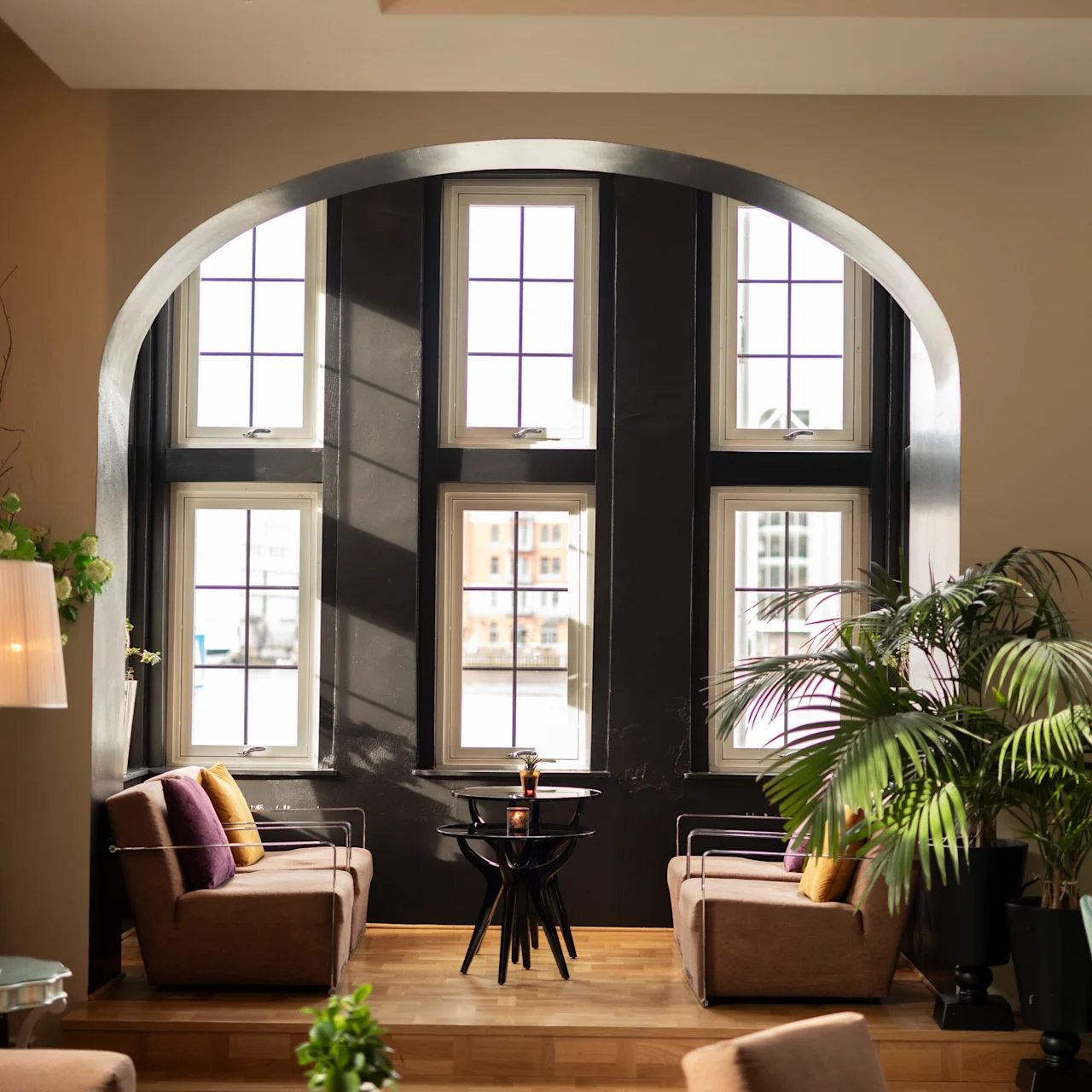 Two plush chairs face each other by a windowed archway, separated by a small table, within a warmly lit room adorned with plants and a tall lamp.