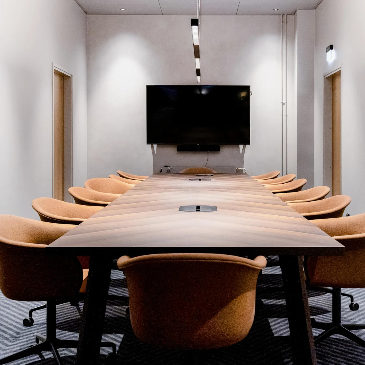 A long conference table surrounded by orange chairs, facing a large wall-mounted television. The room has white walls, a striped carpet, and minimalistic lighting.