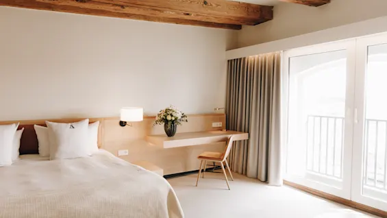 Ljust hotellrum med en stor säng, synliga takbjälkar i trä, ett skrivbord och glasdörrar ut till en balkong.