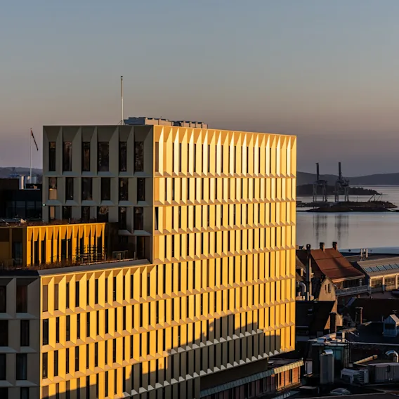 Ilmakuva modernista hotellirakennuksesta, jota kultainen auringonvalo valaisee auringonlaskun aikaan, taustalla kaupunki ja tyyni vuono.