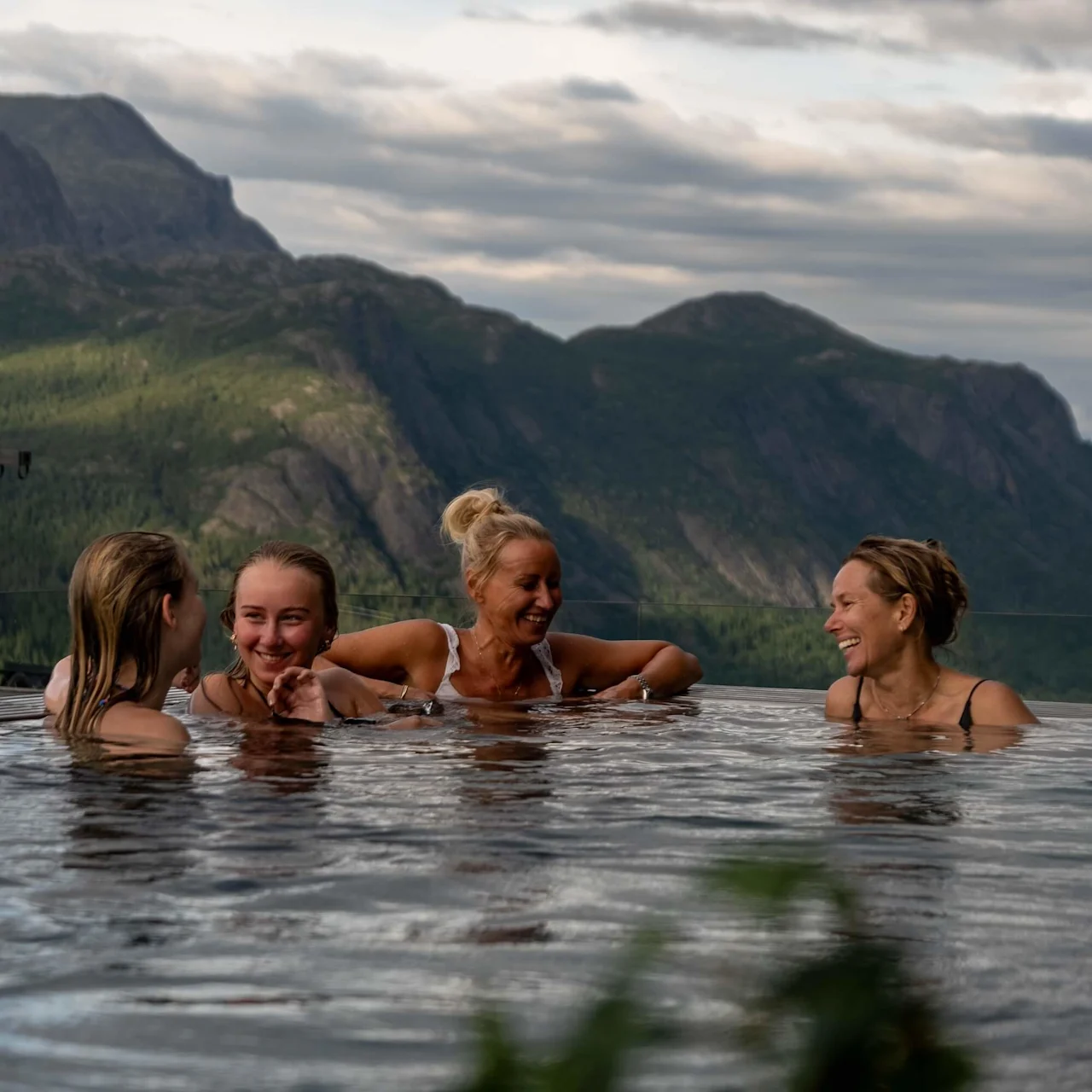 Four women relax and smile in an infinity pool overlooking majestic green mountains and a cloudy sky, creating a serene and joyful atmosphere.
