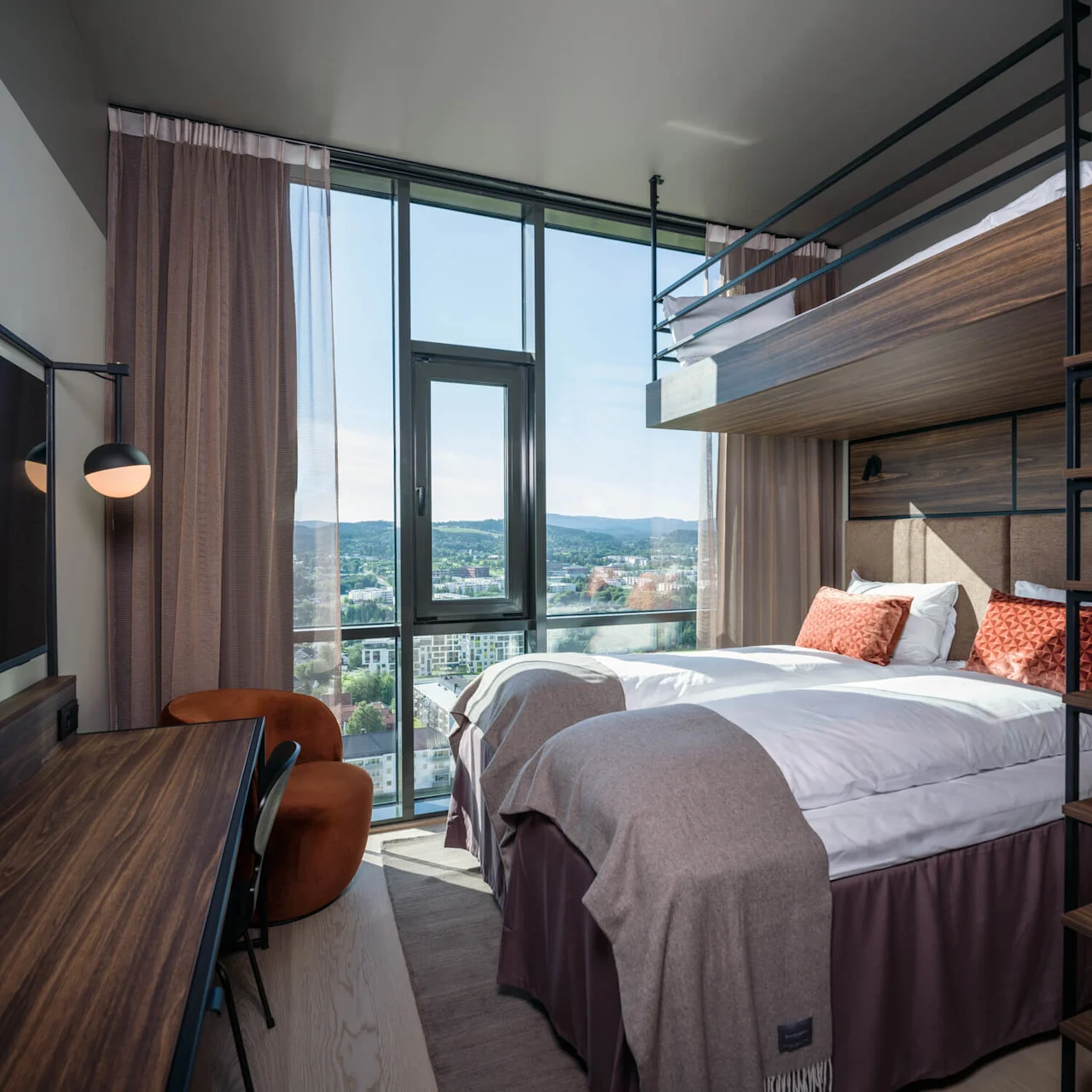 Et moderne hotellrom inneholder en dobbeltseng med oransje puter, en køyeseng ovenfor, et skrivebord med en stol, og store vinduer med utsikt over byen.