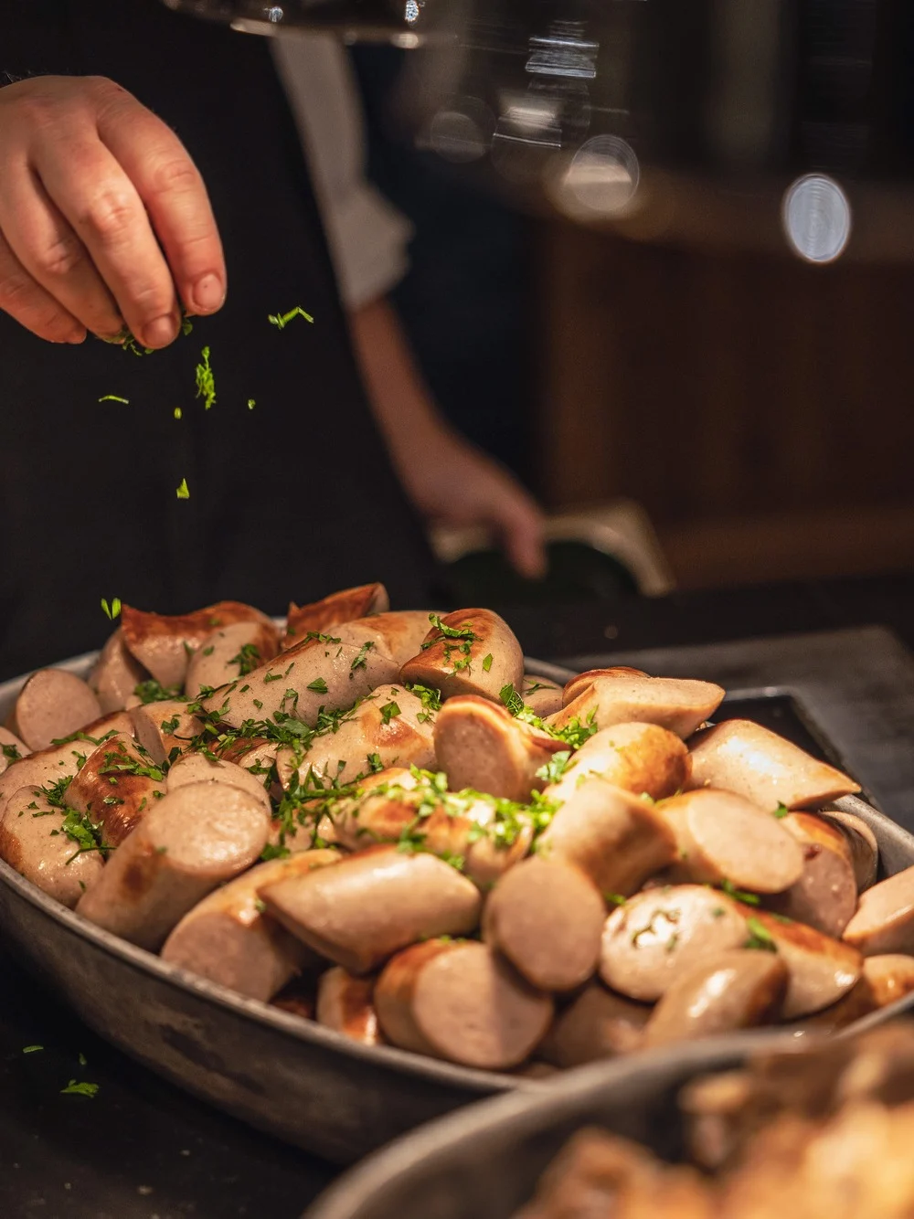 En hånd drysser urter over skivede pølser i en skål, omgitt av andre tilberedte retter på en benk i en varm, svakt opplyst setting.
