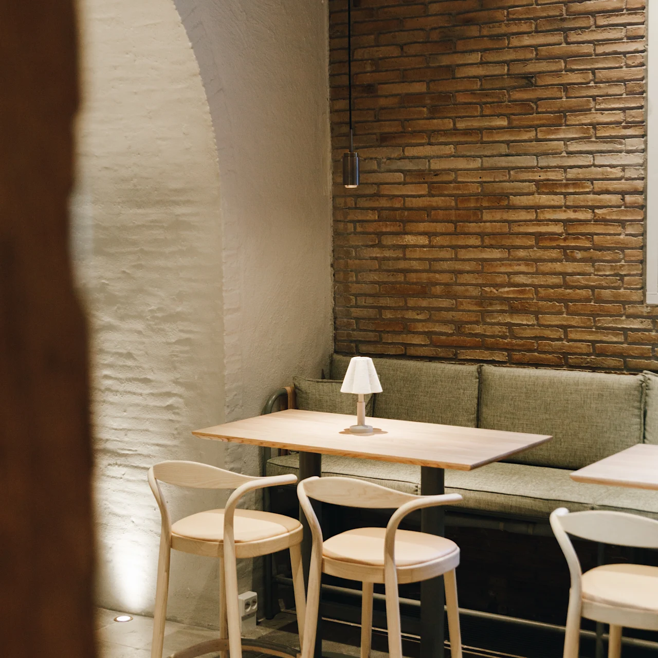 A small lamp sits on a wooden table surrounded by chairs and a cushioned bench, set against a brick and plaster wall in a dimly lit room.