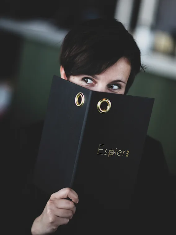 A person peeking over a black menu or binder with the text Espier BAR, looking to the side.