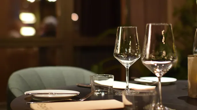A table set with empty wine glasses, plates, and cutlery in a softly lit, elegant restaurant. A large window and blurred lights provide a warm, cozy atmosphere.