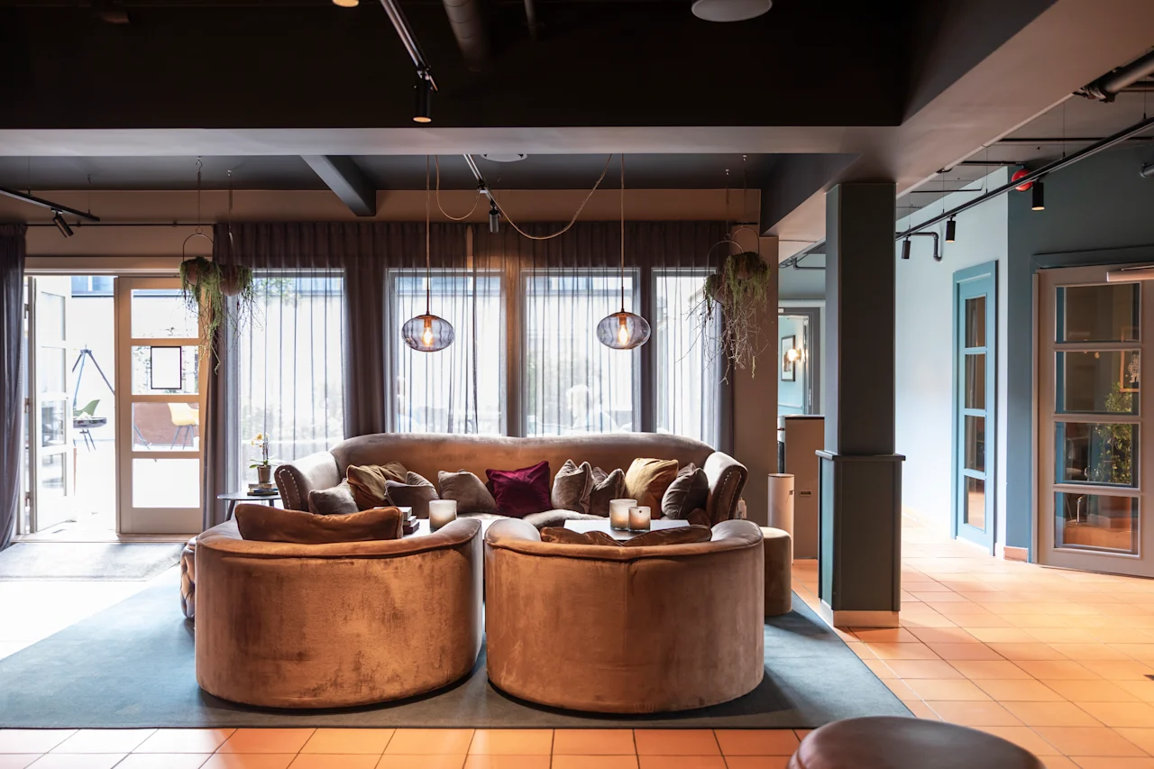 En stor, mjuk hörnsoffa med runda kanter omges av kuddar i en mysig lounge. Mjuk belysning och krukväxter förstärker den moderna, inbjudande interiören.