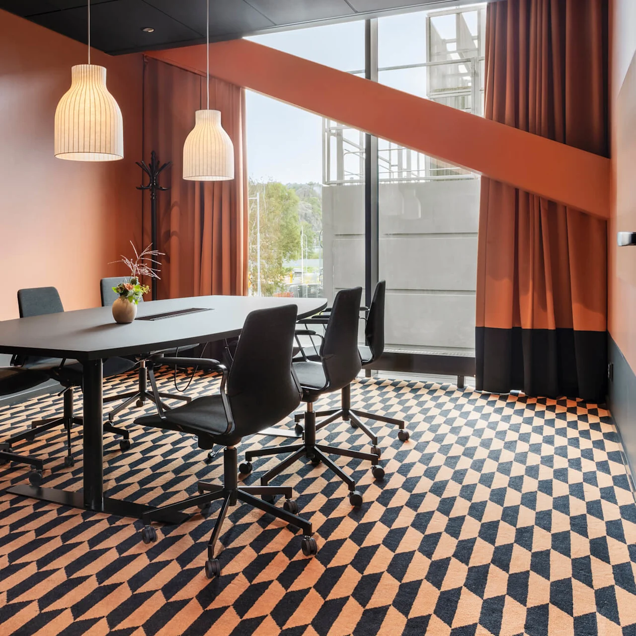 Ett modernt konferensrum med ett svart bord och fyra stolar, hängande lampor och geometrisk matta, omgiven av orange väggar och stort fönster med orange gardiner.