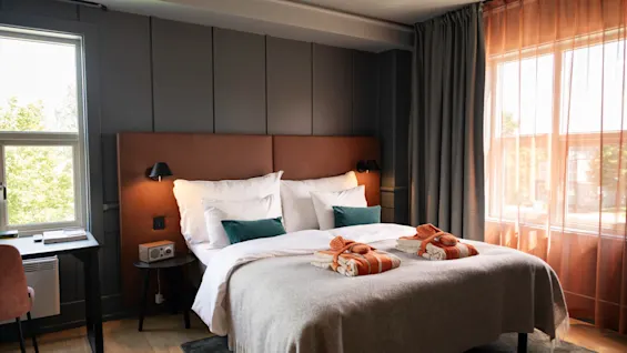 En seng med orange håndklæder og grønne puder står i et moderne hotelværelse med et skrivebord, store vinduer og blødt lys.