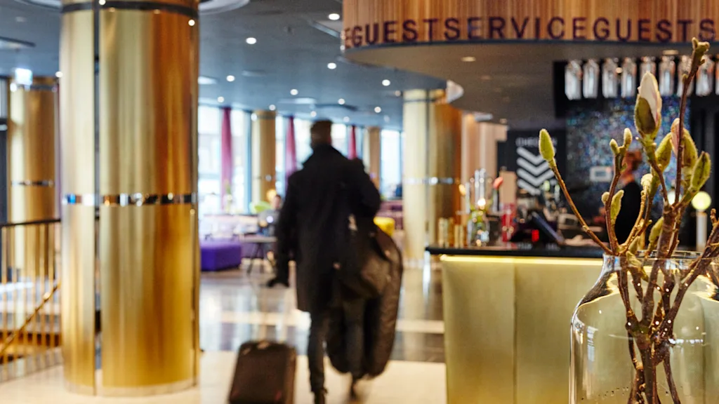 En person går gennem en stilfuld hotellobby og trækker en kuffert. Det moderne rum har gyldne søjler, lilla siddepladser og en reception med blomsterarrangement. Tekst: "GUEST SERVICE".