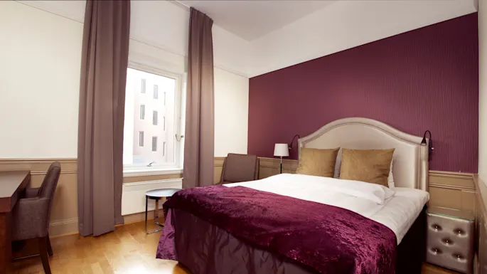 A neatly made bed with a purple throw in a bedroom with a maroon accent wall, window, and furnishings.