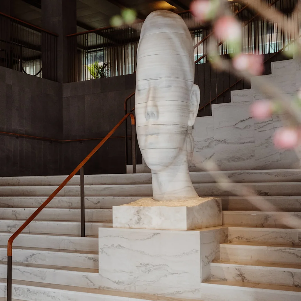 Skulptur av ett fridfullt ansikte på marmorsteg, omgiven av en modern interiör med mörka väggar och räcken. Oskärpa rosa blommor i förgrunden.