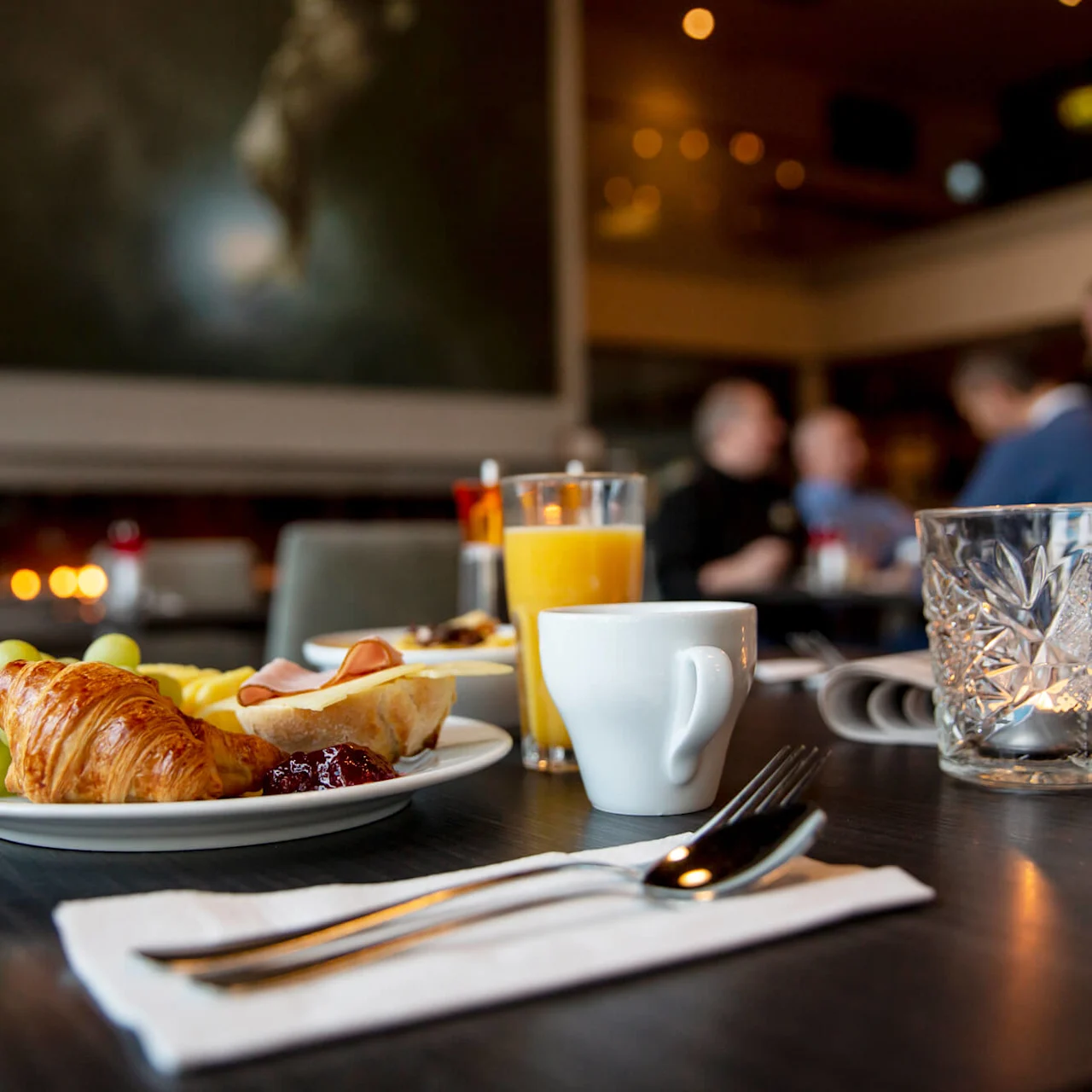 Et frokostbord viser en croissant, druer og en kaffekopp. I nærheten står appelsinjuice på en serviett med bestikk. Folk samtaler i den mykt opplyste, elegante restauranten.