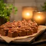Chocolate cake squares with swirled frosting sit on parchment paper atop a wooden board. Two lit candles create a warm glow, surrounded by green potted plants.