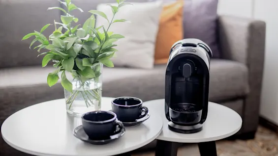 A coffee machine rests on a side table with two dark cups. Nearby, a vase with green plants adds a fresh touch. Background shows a sofa with colorful cushions.