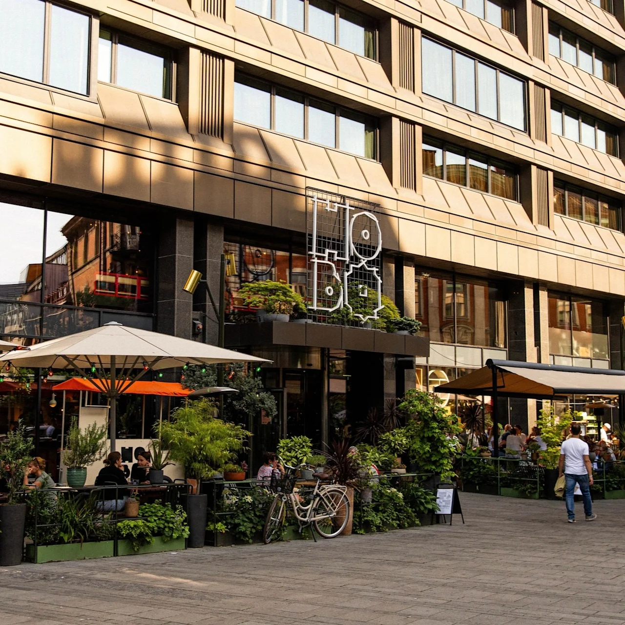 People sitting under large umbrellas at an outdoor café surrounded by plants and bicycles. Large building backdrop with “Hobo” sign on the wall, creating a relaxed urban atmosphere.