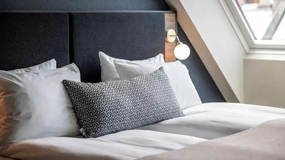 Säng med vita lakan och mönstrad kudde under en modern lampa, bredvid ett lutande fönster i ett elegant sovrum.